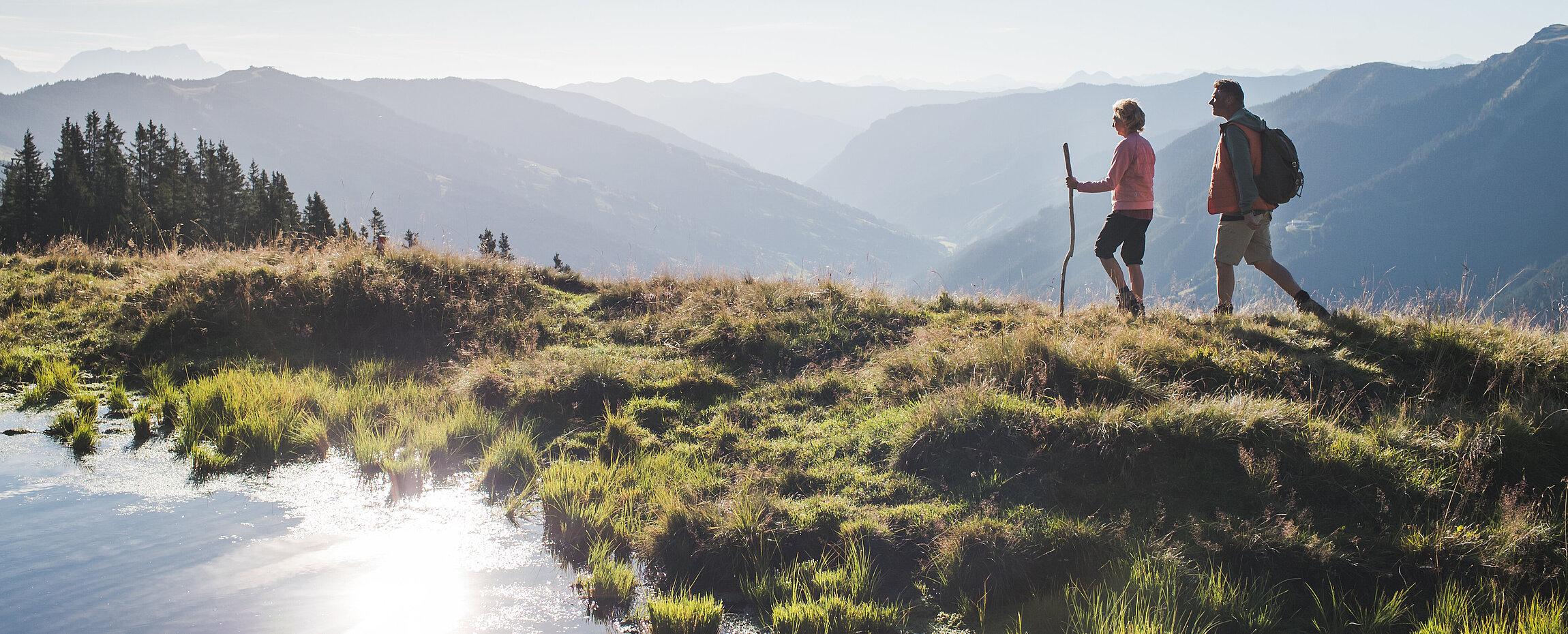 Wanderpaar am Berg mit kleinem Bergteich, dahinter Tallandschaft.