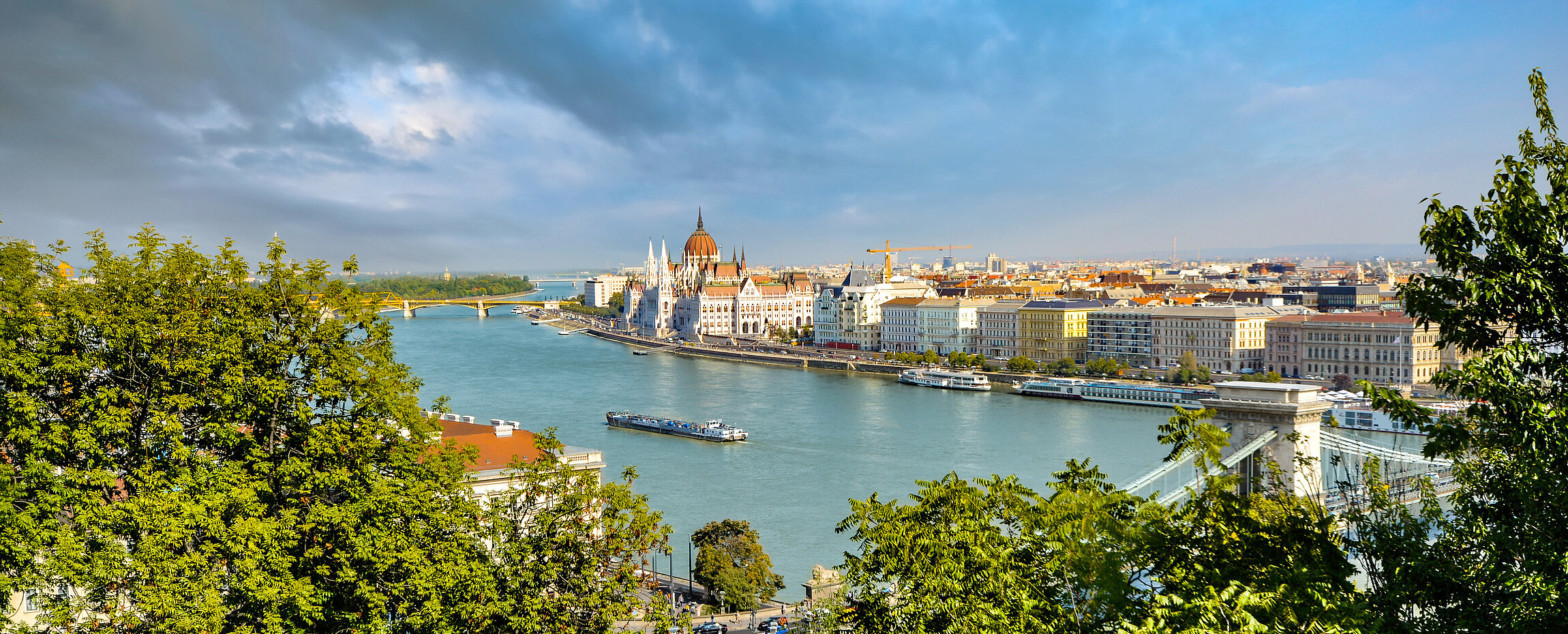 Luftaufnahme der Donau in Budapest mit Blick auf das ungarische Parlamentsgebäude und die Kettenbrücke, umrahmt von grünen Baumwipfeln.