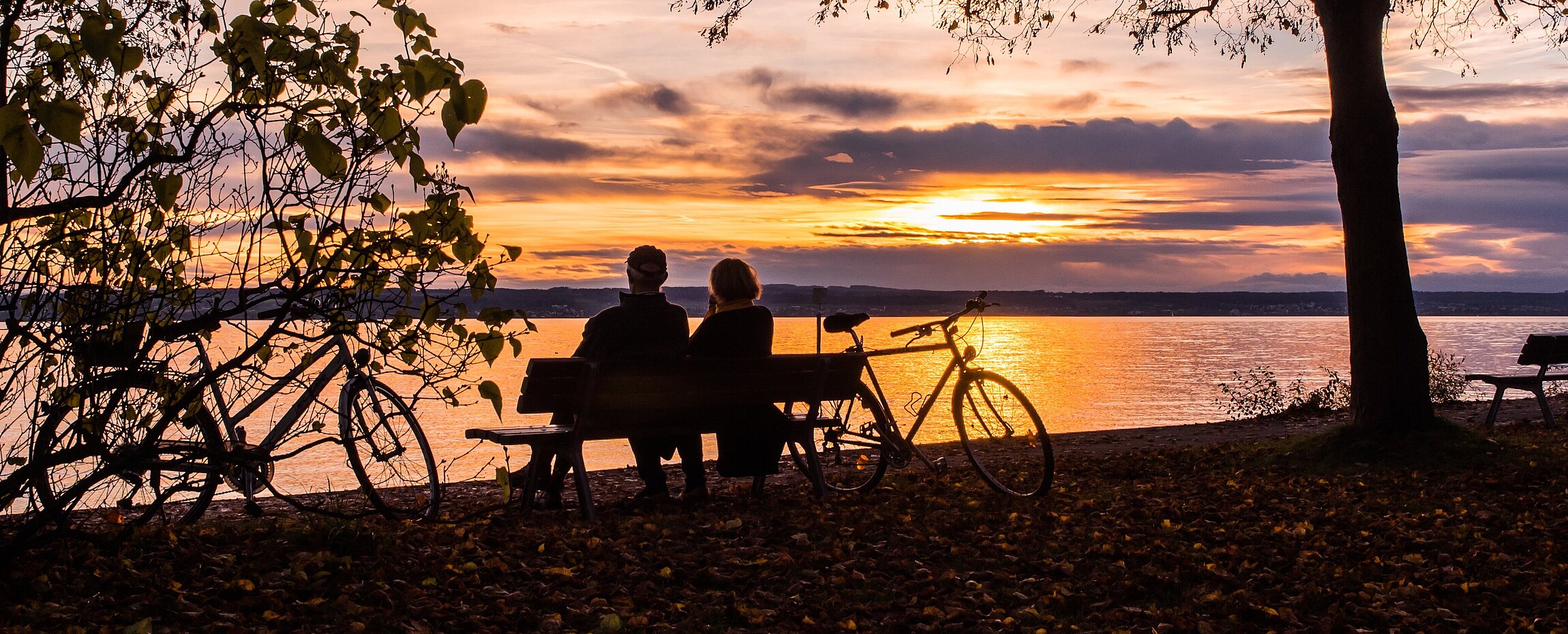 Radfahrer sitzen bei Sonnenuntergang am Ufer des Bodensees auf einer Bank.