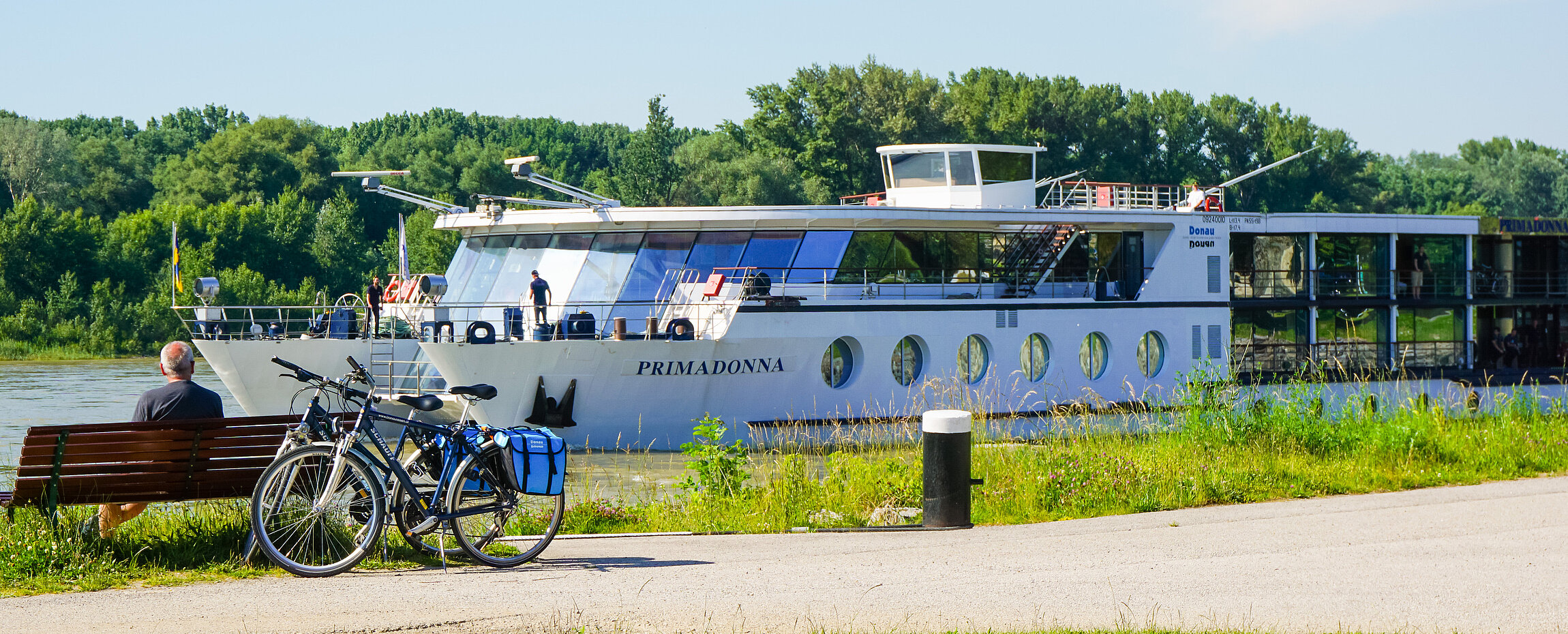 MS Primadonna auf der Donau, während Radfahrer auf einer Bank am Ufer sitzen.