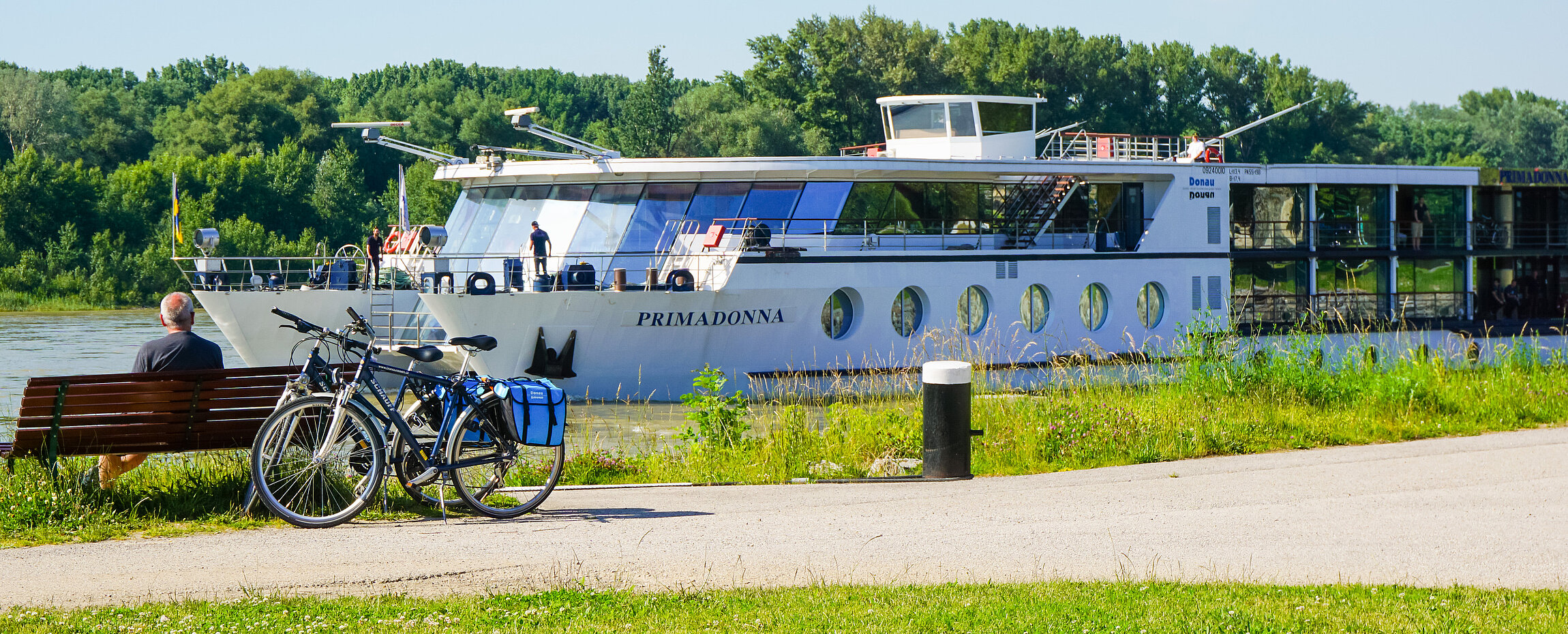 MS Primadonna auf der Donau, während Radfahrer auf einer Bank am Ufer sitzen.