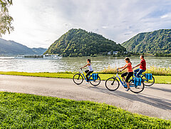 Drei Radfahrer am Donauradweg bei Schlögen, während die MS Primadonna auf der Donau vorbeifährt.
