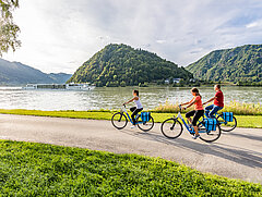 Drei Radfahrer am Donauradweg bei Schlögen, während die MS Primadonna auf der Donau vorbeifährt.